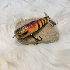 May include: A colorful fishing lure with a black, yellow, and red design. The lure has a curved body, two black hooks, and a small propeller at the end. The lure is resting on a small rock.