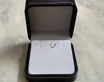 May include: A small, gold-colored nose stud shaped like the letter U, resting inside a dark brown, rectangular jewelry box. The box features a hinged lid and a white, velvet-like interior lining designed to hold small pieces of jewelry securely.