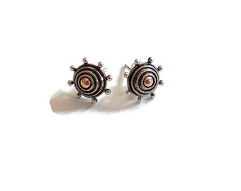 May include: A pair of circular stud earrings featuring concentric silver wire coils and a central polished gold-tone bead. The outer edge of each earring is decorated with small silver spheres, creating a sunburst-like appearance with a metallic finish.