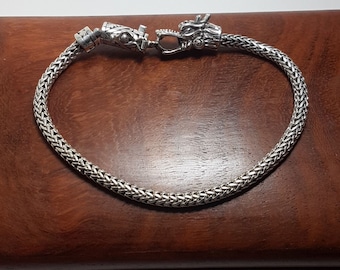 May include: A silver-toned bracelet featuring a woven chain design. The clasp mechanism is shaped like a dragon head, with intricate detailing on the metal surface. The bracelet is circular in shape and displays a metallic sheen throughout the links.