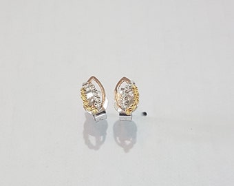 May include: A pair of stud earrings featuring a marquise-shaped frame in polished silver tone. The center of each earring contains a cluster of textured, faceted silver and gold tone elements that create a reflective, multi-dimensional appearance.