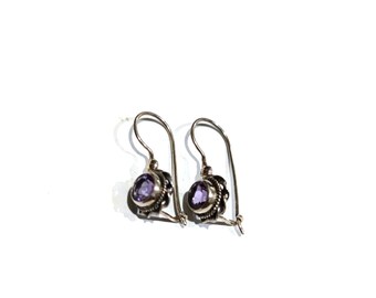 May include: A pair of silver drop earrings featuring round, faceted purple amethyst stones set in a decorative, textured metal bezel. Each earring has a curved wire hook for attachment, showcasing a polished finish and intricate metalwork details.