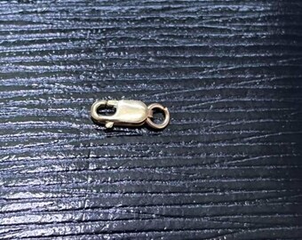 May include: A gold-toned lobster claw clasp featuring a spring-loaded mechanism and a small attached jump ring. The metal component has a smooth, polished finish and is designed for securing jewelry pieces like necklaces or bracelets.