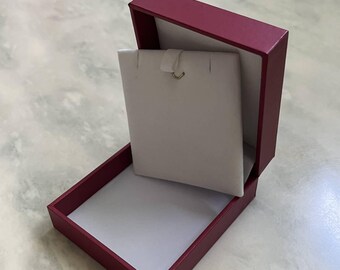 May include: A rectangular jewelry box with a deep burgundy exterior and a white interior. The box features an upright white display card designed for holding a necklace, with a small metal hook visible at the top center of the card.