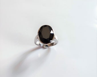 May include: A silver-toned ring featuring a large, oval-shaped, faceted black gemstone set in a smooth metal bezel. The band is polished and reflective, showcasing a clean, metallic finish that complements the dark, central stone.
