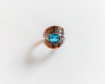 May include: A ring featuring a central oval-cut blue gemstone set within a band of mixed metal tones. The band displays a layered design with a row of small, circular metallic accents bordering the central stone on both the top and bottom edges.