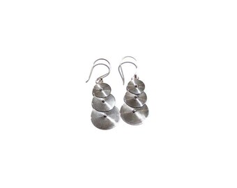 May include: A pair of silver-toned drop earrings featuring three stacked circular discs. Each disc is crafted with a fine, concentric spiral pattern radiating from a central point. The earrings hang from curved silver-toned ear wires.