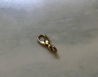 May include: A gold-toned metal lobster claw clasp featuring a spring-loaded mechanism and a small attached jump ring. The jewelry component has a smooth, polished finish and is designed for securing necklaces, bracelets, or other jewelry items.