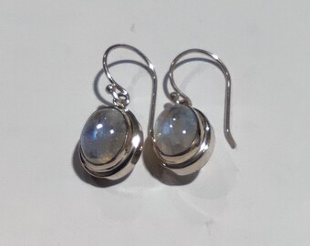May include: A pair of silver drop earrings featuring oval-shaped, translucent moonstones with a subtle blue iridescent sheen. Each stone is set in a smooth, polished silver bezel with a simple curved silver ear wire for hanging the jewelry.