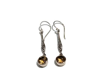 May include: A pair of drop earrings featuring silver-toned metal hooks and decorative stems with intricate filigree patterns. Each earring suspends a single, round, faceted, translucent amber-colored gemstone set in a smooth silver-toned metal base.