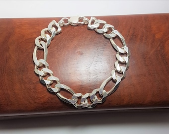 May include: A silver Figaro chain bracelet featuring a repeating pattern of three small circular links followed by one elongated oval link. The polished metal links have a bright, reflective finish and are secured with a lobster claw clasp.