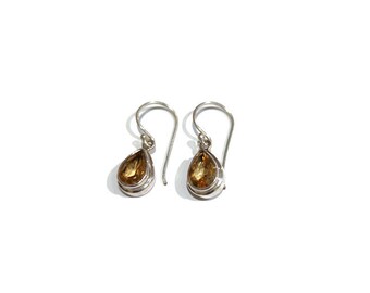 May include: A pair of drop earrings featuring faceted, pear-shaped yellow gemstones set in silver bezels. Each earring hangs from a curved silver ear wire, showcasing a clear, warm-toned stone that reflects light with a polished metallic finish.
