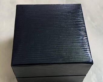 May include: A square, dark navy blue jewelry box with a textured, horizontal lined finish. The box features a hinged lid design with a clean, sharp-edged silhouette, presenting a uniform and solid appearance from a top-down perspective.