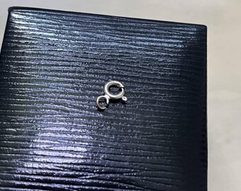 May include: A small, circular silver spring ring clasp featuring a tiny lever mechanism for opening and closing. The metallic jewelry component has a polished, reflective surface and includes a small attached jump ring at the base for secure attachment.