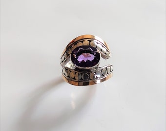 May include: A ring featuring a central oval-cut purple amethyst gemstone. The band is constructed from silver and gold-toned metal, showcasing a decorative pattern of small, raised circular dots along the sides of the setting.