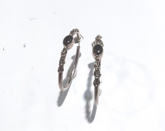 May include: A pair of silver-toned hoop earrings featuring a textured, organic surface design. Each earring is accented with a single, smooth, oval-shaped black stone set near the top post, creating a contrast between the dark stone and metal.