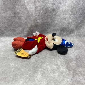 Puede incluir: Un peluche de Mickey Mouse vestido de hechicero, tumbado boca arriba. El juguete lleva una t&uacute;nica de terciopelo rojo con un lazo amarillo, un sombrero azul con estrellas blancas y guantes blancos. El juguete tiene una etiqueta que dice "COLLECTION".