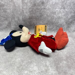 Puede incluir: Un peluche de Mickey Mouse con una chaqueta de terciopelo rojo, guantes blancos y zapatos naranjas. Mickey lleva una capa azul con estrellas blancas y un sombrero negro. Una etiqueta dice "Nostalgic Collection".