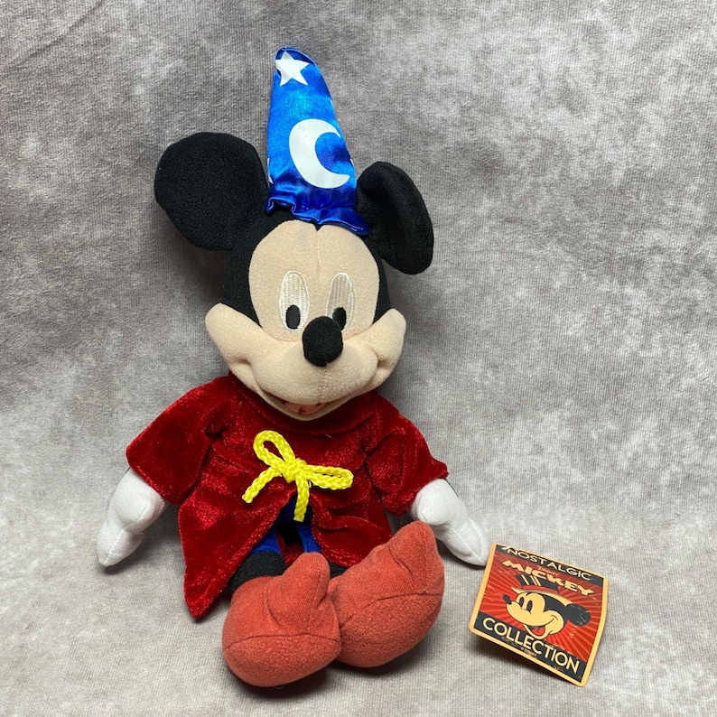 Puede incluir: Un peluche de Mickey Mouse disfrazado de hechicero, con un sombrero azul con una estrella blanca y una luna creciente. Tiene una t&uacute;nica roja con un lazo amarillo y zapatos rojos. Una etiqueta "Nostalgic Mickey Collection" es visible.