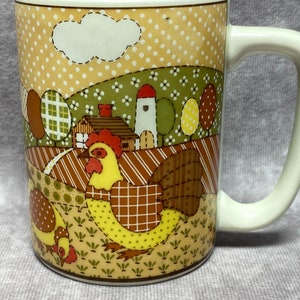 May include: A white ceramic mug with a colourful patchwork design featuring a rooster and a farmhouse scene. The mug has a brown handle.