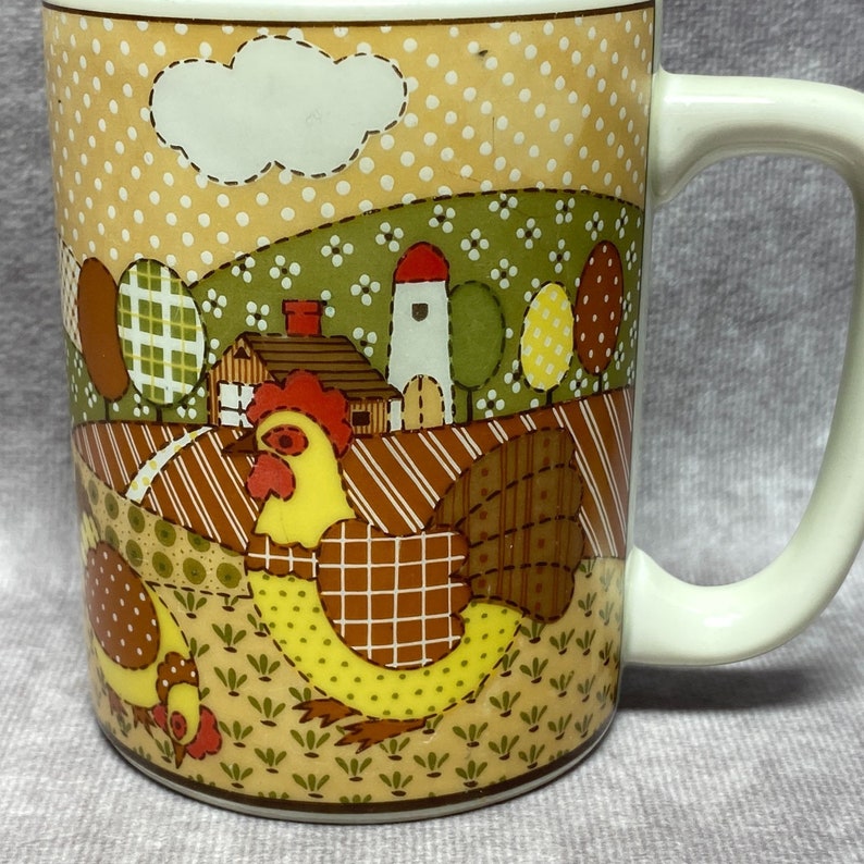 May include: A white ceramic mug with a colourful patchwork design featuring a rooster and a farmhouse scene. The mug has a brown handle.