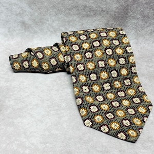 Puede incluir: Una corbata de seda con estampado geométrico verde y marrón. La corbata tiene un fondo blanco con pequeños cuadrados y círculos marrones y naranjas.