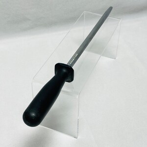 May include: A black-handled steel knife sharpener. The sharpener has a long, slender, silver-colored sharpening rod and a black, ergonomic handle. The sharpener is designed for kitchen use and is displayed on a clear acrylic stand.