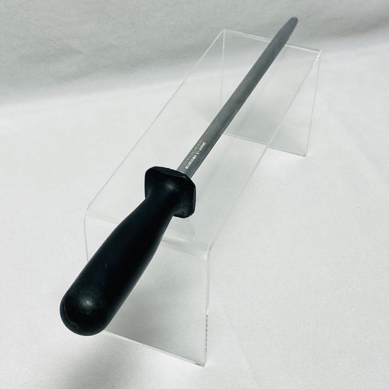 May include: A black-handled steel knife sharpener. The sharpener has a long, slender, silver-colored sharpening rod and a black, ergonomic handle. The sharpener is designed for kitchen use and is displayed on a clear acrylic stand.