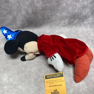 Puede incluir: Un peluche de Mickey Mouse disfrazado de hechicero, con un sombrero azul con estrellas blancas y una t&uacute;nica roja. El juguete tiene orejas negras, una cara beige y guantes blancos. Una etiqueta amarilla con la palabra "NOSTALGIC" est&aacute; adjunta.
