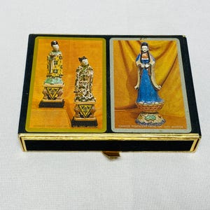 May include: A rectangular black box with gold trim featuring two playing cards on the lid. The left card displays two Chinese porcelain figurines on pedestals. The right card shows a single blue-robed porcelain figure on a decorative base.