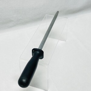 May include: A knife sharpening steel with a black handle and a silver steel rod. The sharpening steel is displayed on a clear acrylic stand against a white background. The handle is rounded and ergonomic.