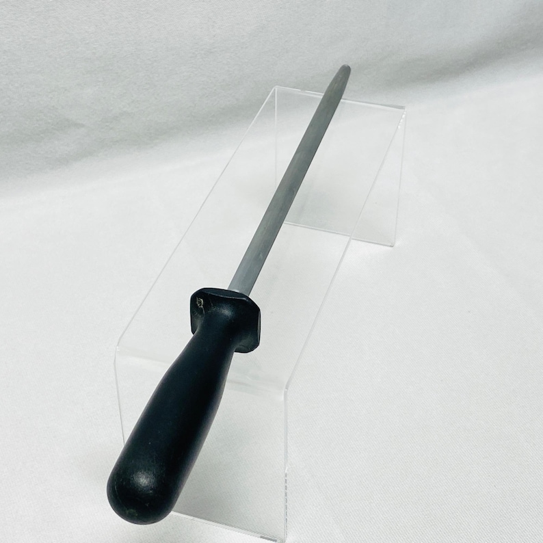 May include: A knife sharpening steel with a black handle and a silver steel rod. The sharpening steel is displayed on a clear acrylic stand against a white background. The handle is rounded and ergonomic.