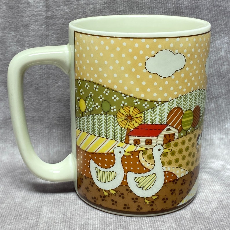 May include: A white ceramic mug with a handle features a patchwork design of a countryside scene with a house, trees, and two geese. The background is a light brown with white polka dots.