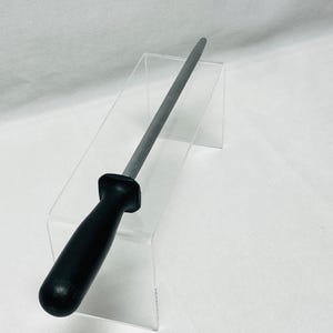 May include: A steel knife sharpening rod with a black handle. The rod is cylindrical and tapers to a point. The handle is made of a smooth, solid black material. The tool is designed for sharpening knives.