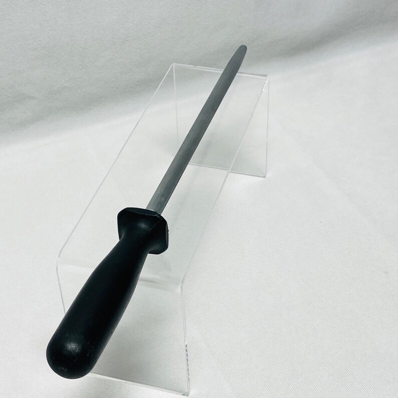 May include: A steel knife sharpening rod with a black handle. The rod is cylindrical and tapers to a point. The handle is made of a smooth, solid black material. The tool is designed for sharpening knives.