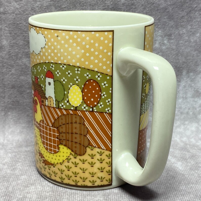 May include: A white ceramic mug with a handle. The mug features a patchwork design with a rooster, a farmhouse, trees, and a field. The colours are brown, yellow, green, and red.