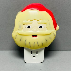 May include: A white plastic nightlight shaped like Santa Claus's head. Santa has a red hat, a white beard, and a smiling face. The nightlight plugs into a standard electrical outlet.