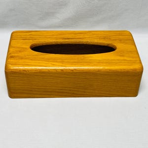 Puede incluir: Un dispensador de pañuelos de madera rectangular con un acabado liso y pulido. La caja es de color marrón dorado cálido y presenta una abertura ovalada larga en la parte superior para dispensar pañuelos. El diseño es simple y clásico.