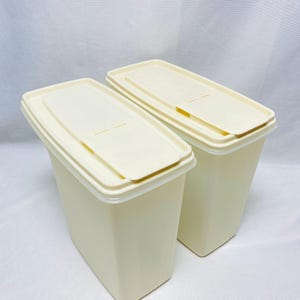 May include: Two rectangular off-white plastic storage containers with matching lids. Each container features a hinged flip-top opening on the lid, allowing for easy access to the contents inside. The containers have a smooth, matte finish.