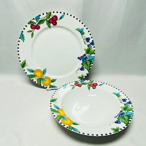 May include: A white ceramic dinner plate and a matching soup bowl, both featuring a black and white checkered rim. The pieces are decorated with colorful illustrations of red cherries, blue grapes, and yellow citrus fruits with green leaves.