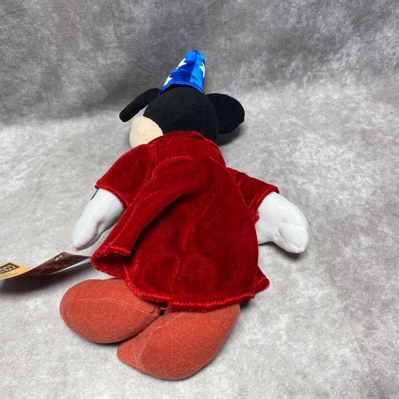 Puede incluir: Un peluche de Mickey Mouse disfrazado de hechicero, con un sombrero azul con estrellas blancas y una t&uacute;nica de terciopelo rojo. El juguete tiene orejas negras, una cara blanca y pies rojos. Una etiqueta est&aacute; adjunta.