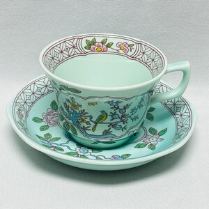 May include: A light mint green porcelain teacup and saucer set featuring a delicate hand-painted design of a small yellow and blue bird perched on a flowering branch, accented by a geometric border pattern in dark red and white along the rim.