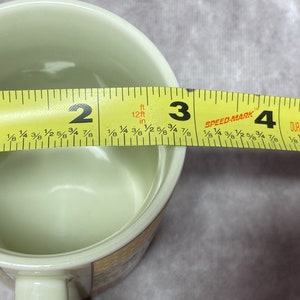 May include: A white ceramic mug with a floral design. The mug is being measured with a yellow tape measure, showing a diameter of 3 inches.