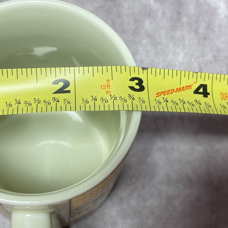 May include: A white ceramic mug with a floral design. The mug is being measured with a yellow tape measure, showing a diameter of 3 inches.