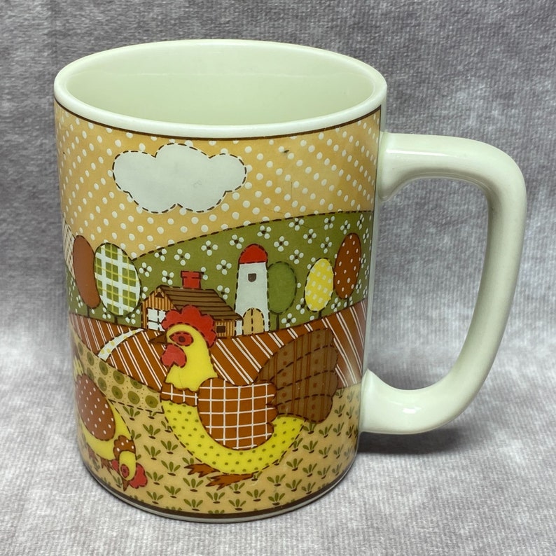 May include: A white ceramic mug with a brown, yellow and green patchwork design featuring a rooster and a farmhouse. The mug has a white handle.