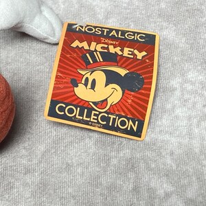 Puede incluir: Una etiqueta rectangular con el texto "NOSTALGIC MICKEY COLLECTION" y una imagen de dibujos animados de Mickey Mouse con un sombrero de copa. La etiqueta tiene un fondo rojo y naranja con un dise&ntilde;o radial. La etiqueta est&aacute; sobre una superficie gris.