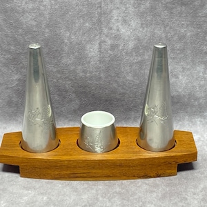 Puede incluir: Un juego de saleros y pimenteros de plata con un soporte para condimentos de cerámica blanca. Los saleros y el soporte están grabados con un diseño de velero y se muestran en una bandeja de madera.