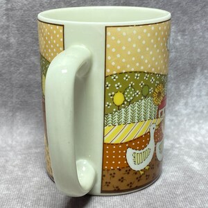 May include: A white ceramic mug with a patchwork design featuring a country scene with geese, a red barn, and yellow and green trees. The mug has a white handle.
