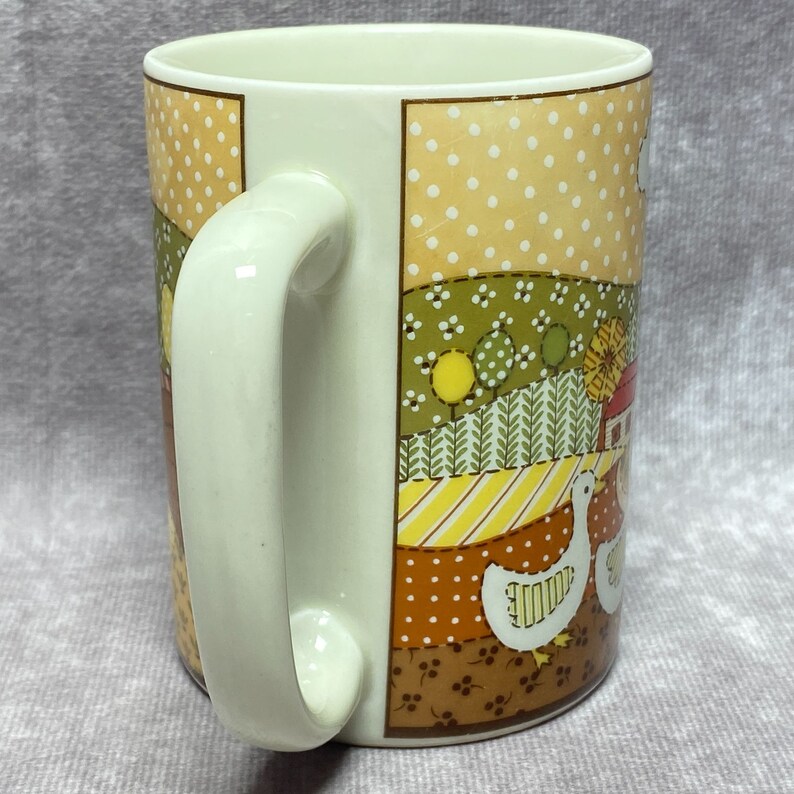 May include: A white ceramic mug with a patchwork design featuring a country scene with geese, a red barn, and yellow and green trees. The mug has a white handle.