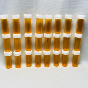 Puede incluir: Una colección de frascos de medicamentos naranjas con tapas blancas. Los frascos están dispuestos en un patrón de cuadrícula, apilados uno encima del otro. El fondo es una superficie blanca lisa.